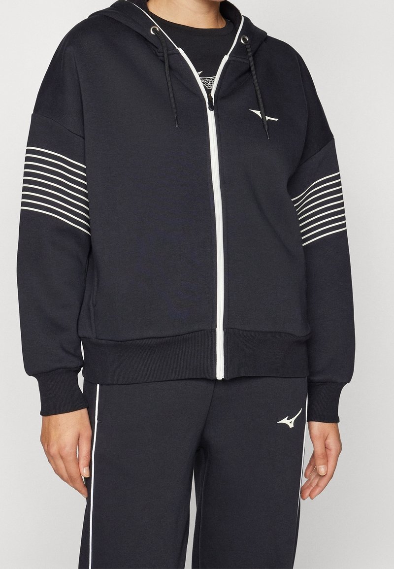 Personne portant un sweat à capuche noir zippé avec des bandes blanches sur les manches et un pantalon noir assorti avec des bandes blanches et un logo.
