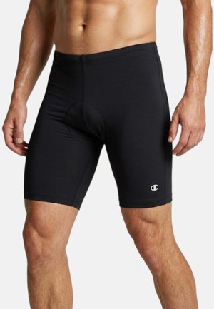 Uomo muscoloso che indossa pantaloncini da ciclismo neri aderenti con un piccolo logo sulla coscia sinistra su uno sfondo semplice.