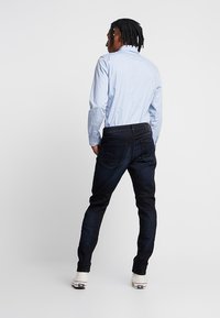 Hombre con rastas vestido con una camisa azul claro, jeans oscuros y zapatillas blancas, de pie de espaldas contra un fondo simple.