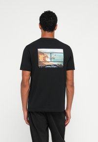 Sort bomulds t-shirt med et grafisk motiv af to kystlandskabsbilleder på ryggen, med "Tommy Hilfiger" tekst og etableringsdato.