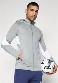 Grå zip-up hættejakke med hvide detaljer, hætte og lange ærmer. Materialet ser blødt og struktureret ud. Modellen holder en sort-hvid fodbold.