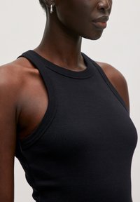 Schwarzes, geripptes Tanktop mit hohem Halsauschnitt und weiten Armlöchern, das eine glatte Textur und eine enganliegende Passform zeigt. Sichtbare silberne Creolen.