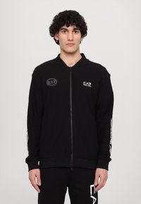 Black zip-up jacket in textured fabric. Features EA7 logo on the chest, contrasting white accents on sleeves, and ribbed cuffs.