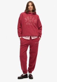 Bordeaux oversized hoodie met logo, voorzak en bijpassende sweatpants. Gladde textuur, ribgebreide boorden en elastische tailleband. Zwarte schoenen.
