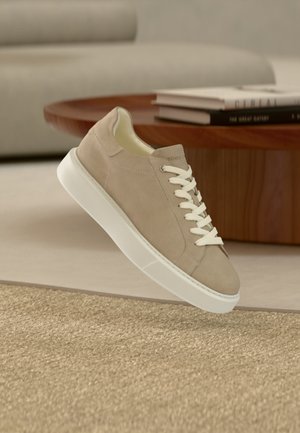 Beżowy zamszowy sneaker z białymi sznurowadłami i grubą białą podeszwą unoszący się nad teksturowanym beżowym dywanem w nowoczesnym salonie.