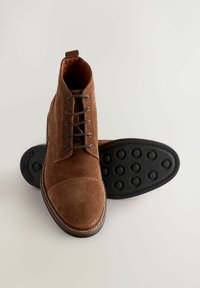 Braune Wildleder-Stiefeletten mit Schnürung und einer strukturierten schwarzen Sohle, von oben auf einem hellen Hintergrund präsentiert.