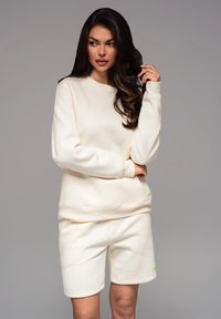 Completo in morbido tessuto composto da felpa color crema e pantaloni corti. La felpa ha un collo rotondo e polsini a costine; i pantaloni corti hanno un elastico in vita.
