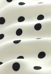 Tissu blanc à pois noirs uniformément espacés, présenté avec de légers plis mettant en valeur une texture lisse.