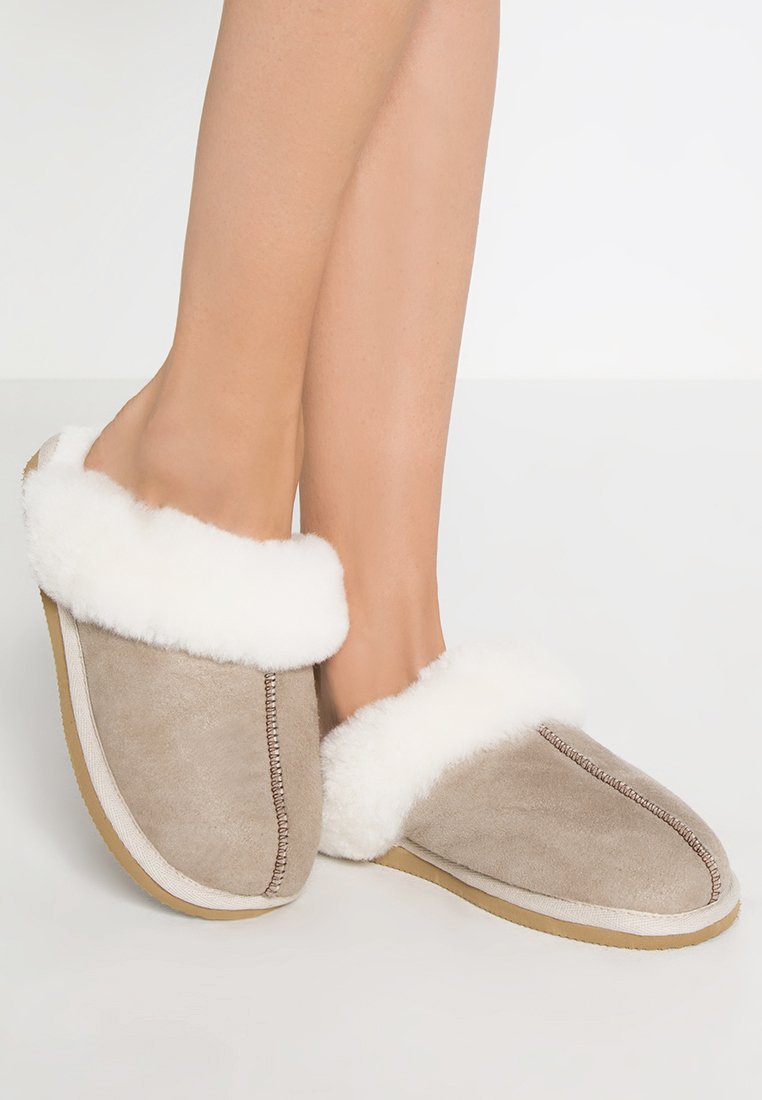 Ciabatte in camoscio beige con polsino in pelliccia sintetica bianca, dettagli cuciti e una suola morbida. Design con tacco aperto e interno soffice per il massimo del comfort.
