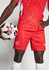 Piros atlétikai mez fehér részletekkel és texturált mintával, hozzáillő piros rövidnadrággal, mely logót visel. A játékos fekete-fehér focilabdát tart.