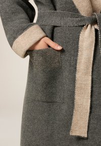 Graue Wollrobe mit einem beigen Kragen und Gürtel. Verfügt über eine Tasche mit metallischen Verzierungen, die einen Texturkontrast und elegantes Design zeigen.