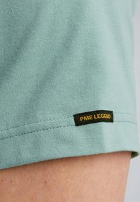 Tela de camiseta verde menta con una textura suave; presenta una etiqueta negra con "PME LEGEND" en costuras amarillas en el dobladillo.