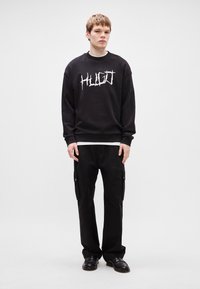 Μαύρη μπλούζα με γραφικό "HUGO" σε άσπρο, συνδυασμένη με μαύρο cargo παντελόνι που διαθέτει πλαϊνές τσέπες, φορεμένη με μαύρα παπούτσια.