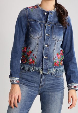 Denimjakke med slidte kanter og farverig blomsterbroderi, båret med matchende blå jeans, hænder afslappet ved siderne.
