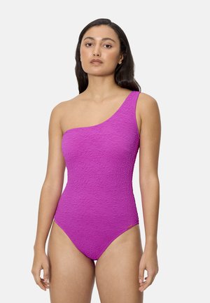 Mujer con un bañador texturizado de un solo hombro en color rosa brillante, de pie con los brazos relajados contra un fondo liso de color gris claro.