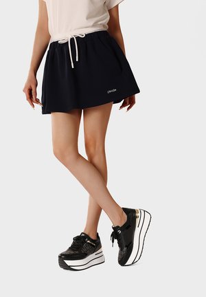 Gambe incrociate con una minigonna nera con coulisse e sneakers platform nere con suola bianca e dettagli patternati.