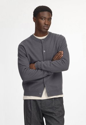 Giovane uomo con le braccia incrociate che indossa un cardigan grigio abbottonato sopra una camicia bianca e pantaloni grigi, in piedi davanti a uno sfondo bianco semplice.