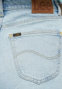 Jasnoniebieskie dżinsy z tylną kieszenią, ozdobioną żółtymi szwami i małą czarną metką z napisem "Lee". Brązowa skórzana metka nad kieszenią.