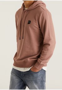 Sudadera marrón de material suave y acanalado con un bolsillo tipo canguro, capucha con cordón y un pequeño logo negro en el pecho. Combinada con jeans claros.