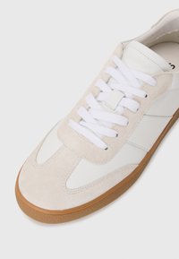 Sneaker bianca con dettagli in suede beige, dotata di lacci bianchi piatti, suola in gomma e dettagli di cucitura laterale. Design semplice e moderno.