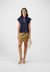 Blusa blu navy a maniche corte con scollatura a forma di chiave, abbinata a una gonna khaki con spacco laterale e sandali bianchi con tacco.