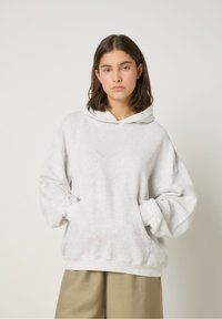 Sudadera gris de gran tamaño hecha de un tejido suave y texturizado, con capucha ajustable y bolsillo tipo canguro delantero. Combinada con pantalones marrón claro.