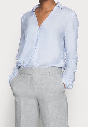 Personne portant une chemise bleu clair à manches longues boutonnée rentrée dans un pantalon taille haute gris clair, les mains dans les poches.