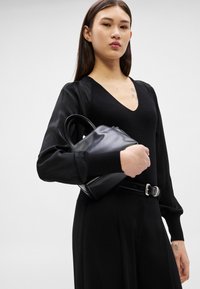 Sieviete melnā V-veida kakla topā un biksēs ar nelielu melnu ādas rokassomiņu zem rokas pret vienkāršu gaišu fonu.