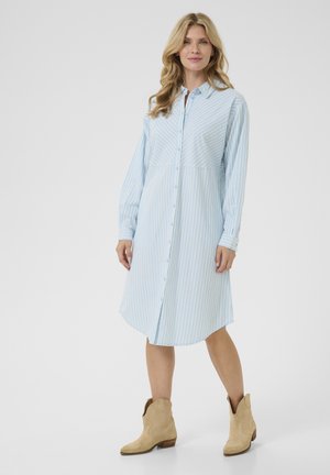 Abito camicia a righe blu chiaro con maniche lunghe, apertura con bottoni e un leggero orlo asimmetrico, abbinato a stivali da cowgirl color sabbia.