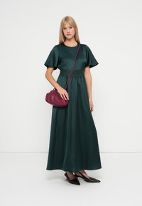 Μακρύ, σκούρο πράσινο σατέν φόρεμα με κοντά μανίκια, στενή μέση και δαντελένια λεπτομέρεια. Συνδυάζεται με μια μπορντό τσάντα χιαστί και μαύρες γόβες.