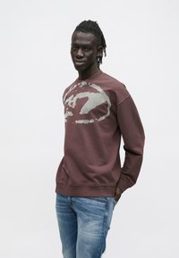 Krvavo rdeč pulover z velikim sivim graphicem v stilu staranja, z okroglim izrezom in rebrastimi manšetami, kombiniran s svetlo modrimi jeans hlačami. Mehka, udobna tkanina.