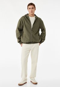Chaqueta con capucha en verde oliva, con cremallera frontal, dobladillo elástico y bolsillos laterales, combinada con pantalones color crema y zapatillas blancas.