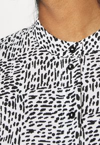 Camicia bianca e nera con colletto a fascetta; il tessuto presenta un motivo irregolare e testurizzato con linee audaci e a contrasto.