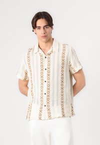 Cămașă cu mânecă scurtă de culoare crem, cu un model geometric brun, cu închidere prin butoni și material texturat. Croială lejeră cu un guler relaxat.