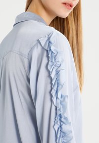 Blouse bleu clair en tissu lisse avec un design à volants le long de la couture de l'épaule et un col classique.