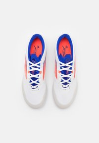 Zapatillas deportivas blancas con acentos en azul y naranja, parte superior texturizada, cordones planos y diseño de punta redondeada. Logo de marca destacado en la plantilla.