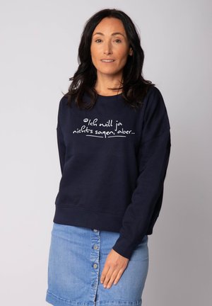 Navy blue sweatshirt with white text, relaxed fit, ribbed cuffs, and hem. Paired with a light denim skirt featuring a button-down front.