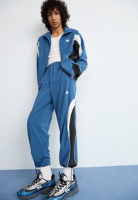 Blue tracksuit featuring black and white accents, with a zip-up jacket and elastic waistband pants. Paired with silver and blue sneakers.