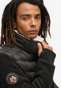 Sort puffjakke med fleeceærmer, høj krave og ribbede manchetter. Har et rundt logo med "Superdry" på venstre ærme.