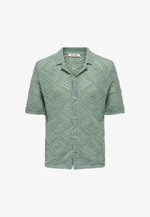 Kratka pletena jopica s kratkimi rokavi v svetlo zeleni barvi, ki ima geometrijski diamantni vzorec, sprednje gumbe in ovratnik.