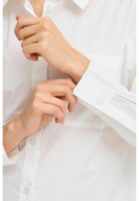 Camicia bianca con colletto, dotata di tasca sul petto, bottoni visibili e maniche arrotolate. Il tessuto sembra liscio e leggero.
