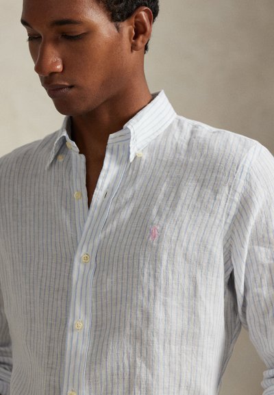 Πουκάμισο με κουμπιά σε ανοιχτό μπλε και λευκές ρίγες, κατασκευασμένο από υφασμάτινη υφή· διαθέτει μικρό ροζ λογότυπο και κίτρινα κουμπιά.
