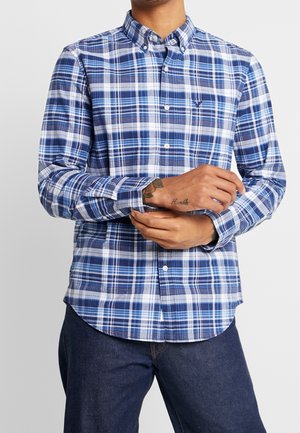 Mężczyzna poprawiający mankiet niebiesko-białej koszuli w kratę, noszący ciemne dżinsy, ukazujący tatuaż "Humble" na lewym nadgarstku.