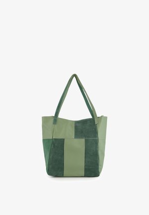 Grøn patchwork-tote-taske med to håndtag, der kombinerer ruskind og glatte læderpaneler, står oprejst på en hvid baggrund.