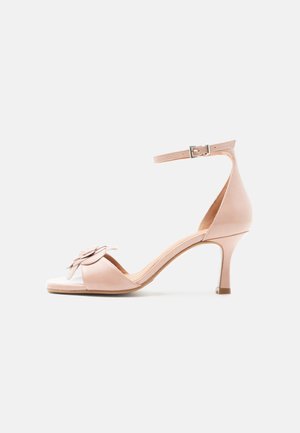 Nøttefarget, åpen tå high heel sandal med ankelstropp, spenne og blomsterdekorasjon på frontstroppen mot hvit bakgrunn.