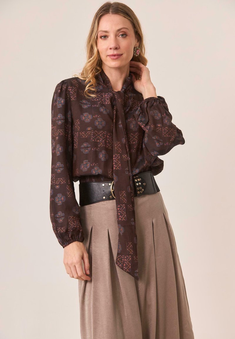 Blusa marrón con estampado, cuello con lazo, mangas largas abullonadas y cintura ceñida con cinturón negro. Complementada con una falda plisada en tono topo.