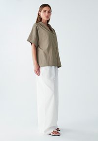 Mujer vistiendo una camisa de botón de cortos de manga oversized en beige, pantalones anchos blancos, sandalias blancas y pendientes de aro dorados sobre fondo blanco.