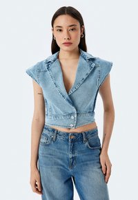 Gilet in denim azzurro chiaro, cropped, con scollatura a V profonda, spalline e bottoni frontali, abbinato a jeans blu a vita alta.