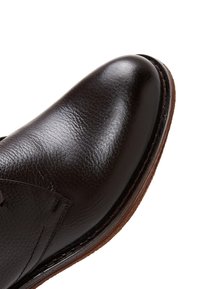 Chaussure en cuir marron avec une surface lisse et polie, un bout légèrement pointu et des coutures contrastantes le long d'une semelle texturée.