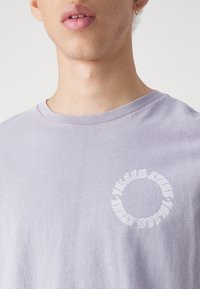 Camiseta de algodón gris-púrpura con un gráfico redondo en blanco. Manga corta, cuello redondo y una textura suave y lisa.